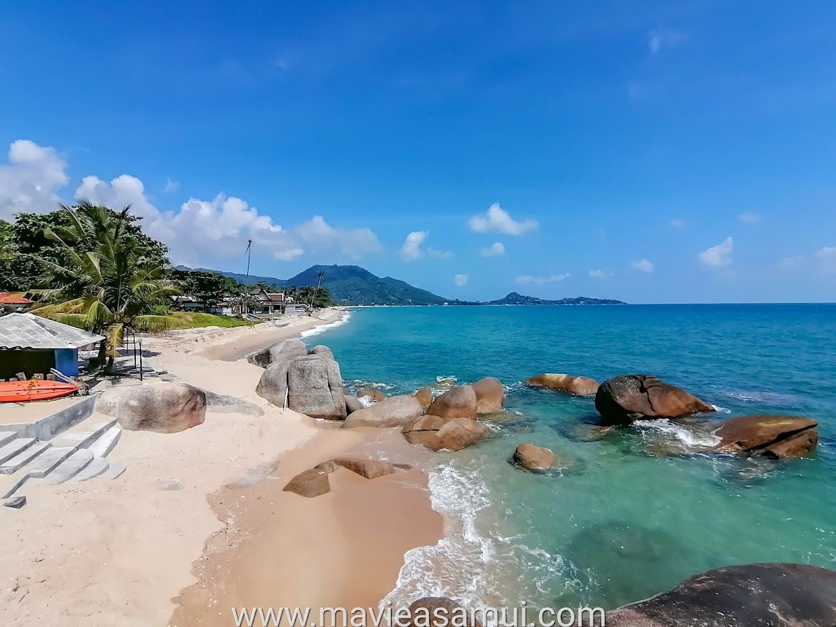 Lamai Beach, Koh Samui — sandy shoreline with large granite boulders at the water's edge, turquoise water, and green hills in the distance
