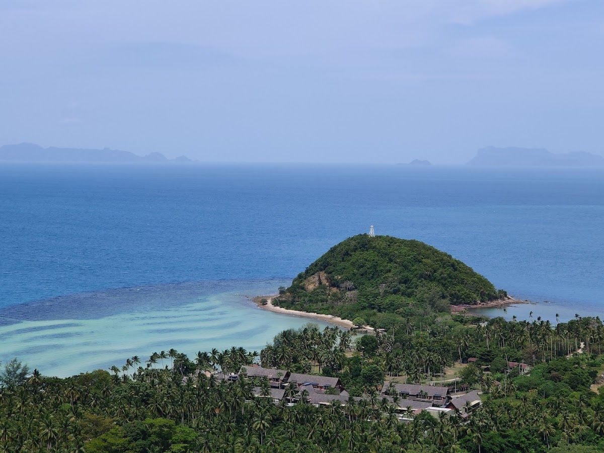 View from a Koh Samui ring road viewpoint showing a small palm-covered island with a lighthouse and turquoise water, with more islands visible through haze in the background