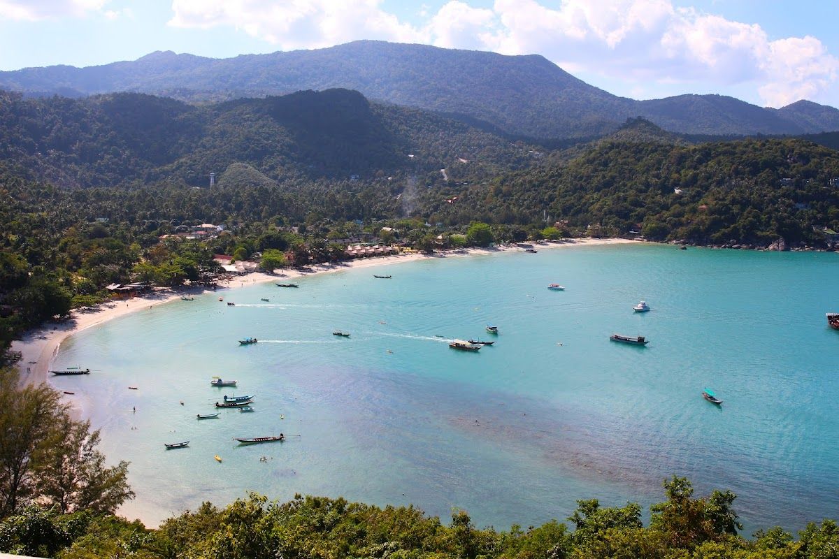 Thong Nai Pan Bay aerial view showing turquoise water, long-tail boats, white sand beach, and jungle-covered hills on Koh Phangan