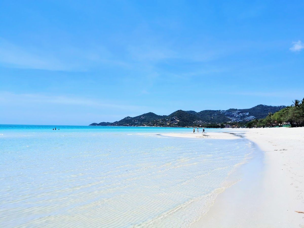 Chaweng Beach, Koh Samui, with white sand, clear turquoise water, and forested hills in the background on a sunny day